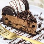 An overhead shot shows a big slice of chocolate Oreo cheesecake on a white rimmed plate that is drizzled with a chocolate sauce. The slice of cheesecake is covered in more crushed Oreos, whipped cream and a chocolate drizzle.