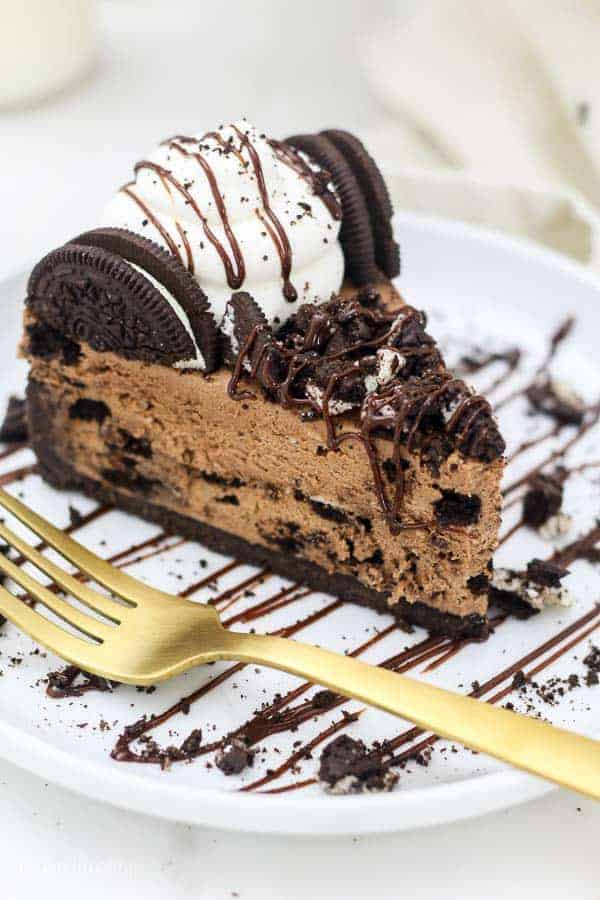 An overhead shot shows a big slice of chocolate Oreo cheesecake on a white rimmed plate that is drizzled with a chocolate sauce. The slice of cheesecake is covered in more crushed Oreos, whipped cream and a chocolate drizzle.