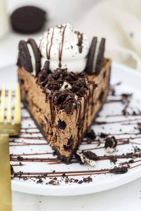 A head on shot of a slice of cheesecake shows all the crumbled Oreos on top with a chocolate drizzle