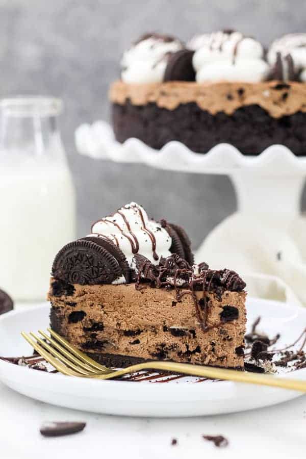 A gold fork on a white rimmed plate is laying next to a big slice of chocolate Oreo cheesecake it covered in crushed Oreos.