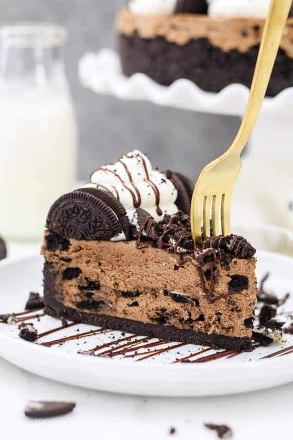 A gold fork is sinking into a slice of chocolate Oreo cheesecake sitting on a white rimmed plate with a chocolate glaze drizzle.