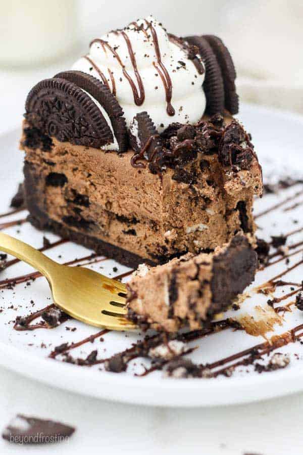 A gold fork with a bite of of cheesecake on it, the slice has a few bites missing and it's garnished with chocolate drizzle, more Oreos and a big dollop of whipped cream.