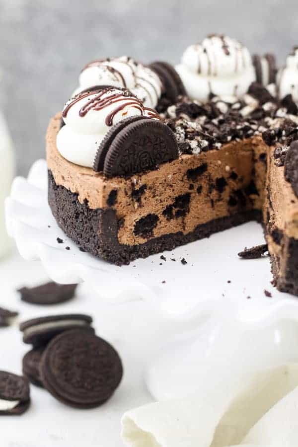 A big slice cut out of a whole chocolate Oreo cheesecake. The cheescake is garnished with whipped cream and more Oreos