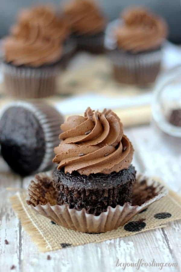 A chocolate cupcake frosted with a big swirl of chocolate frosting, the cupcake is unwrapped sitting on a black polka dot piece of burlap