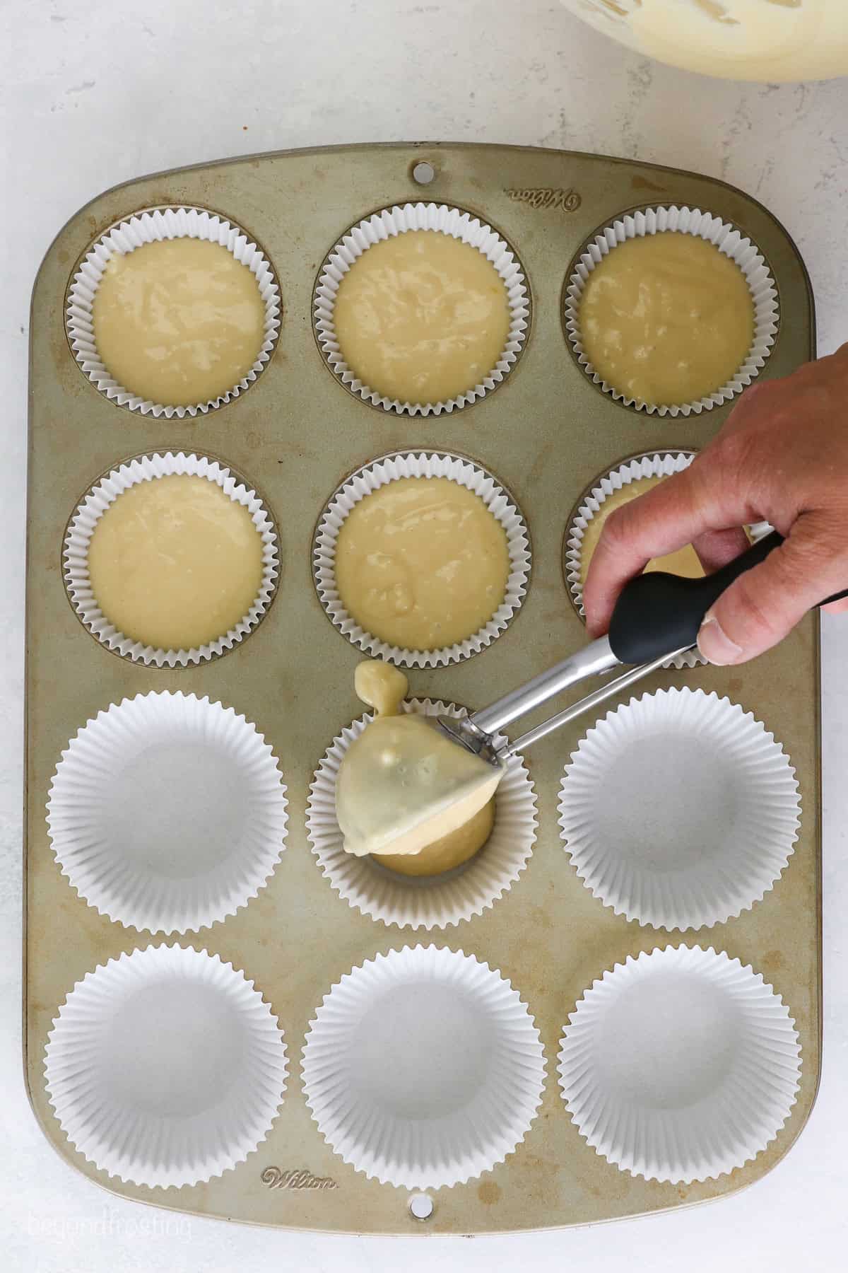 Overhead view of a hand using a cookie scoop to portion yellow cupcake batter in the lined wells of a cupcake pan.