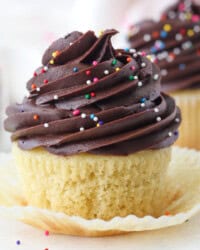 Close up of a yellow cupcake decorated with chocolate frosting and sprinkles in an open cupcake wrapper, with more cupcakes in the background.