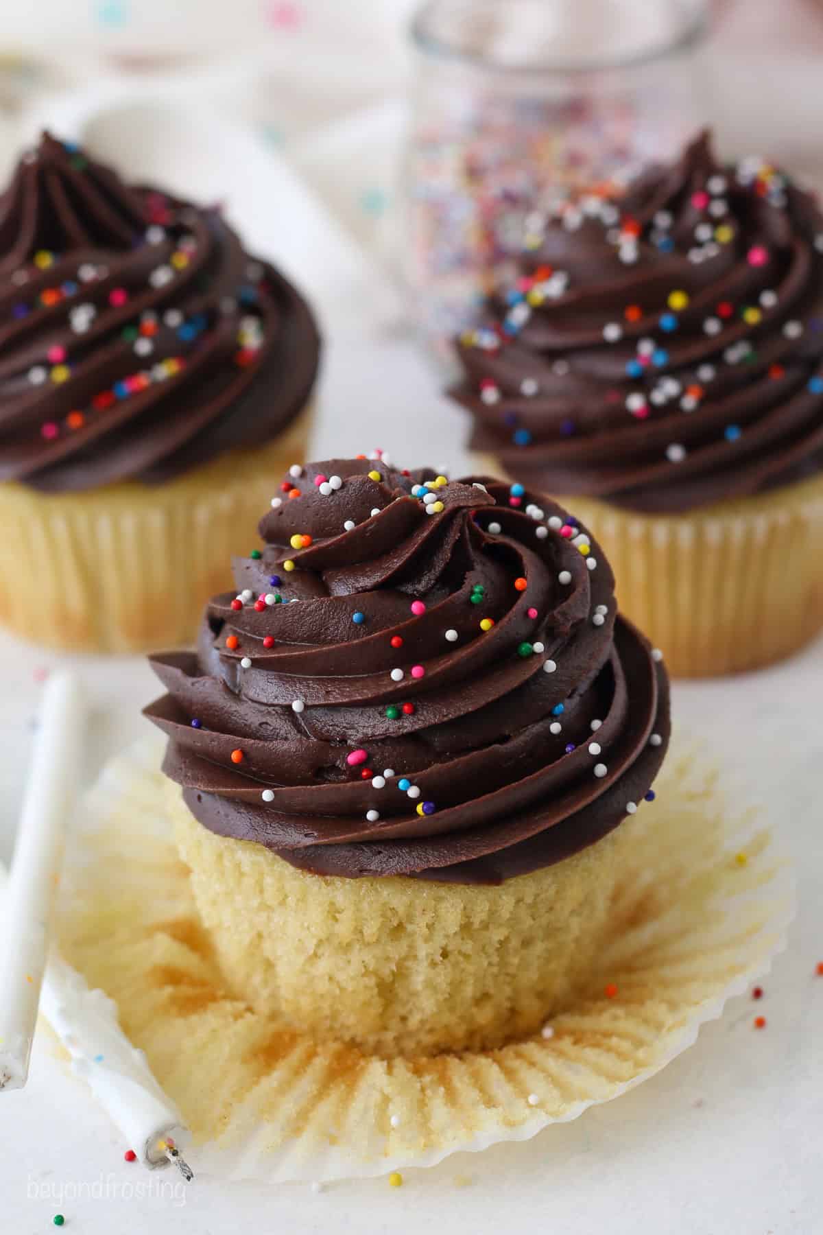 Close up of a yellow cupcake decorated with chocolate frosting and sprinkles in an open cupcake wrapper, with more cupcakes in the background.