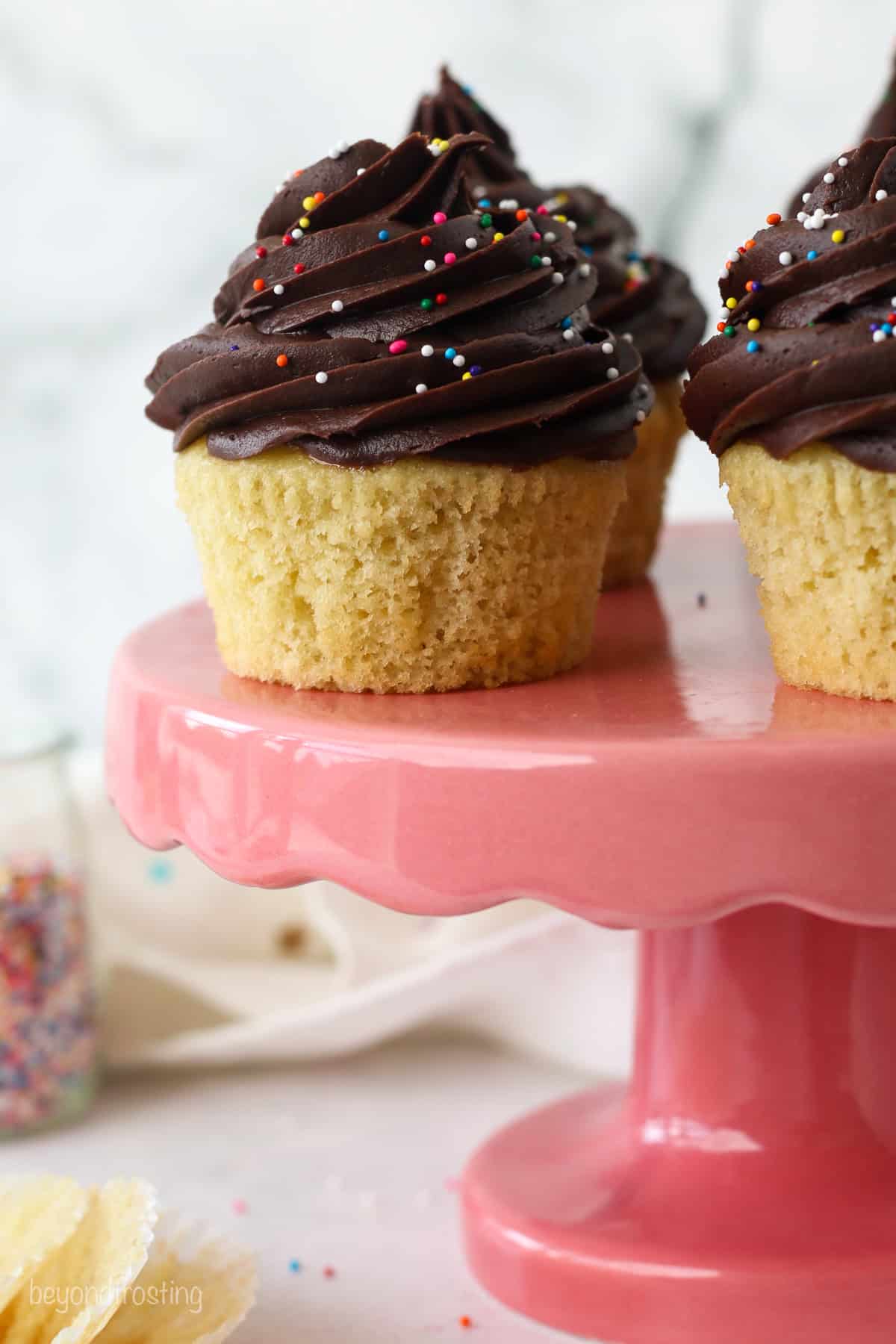 Close up of yellow cupcakes topped with chocolate frosting swirls on a pink cake stand.