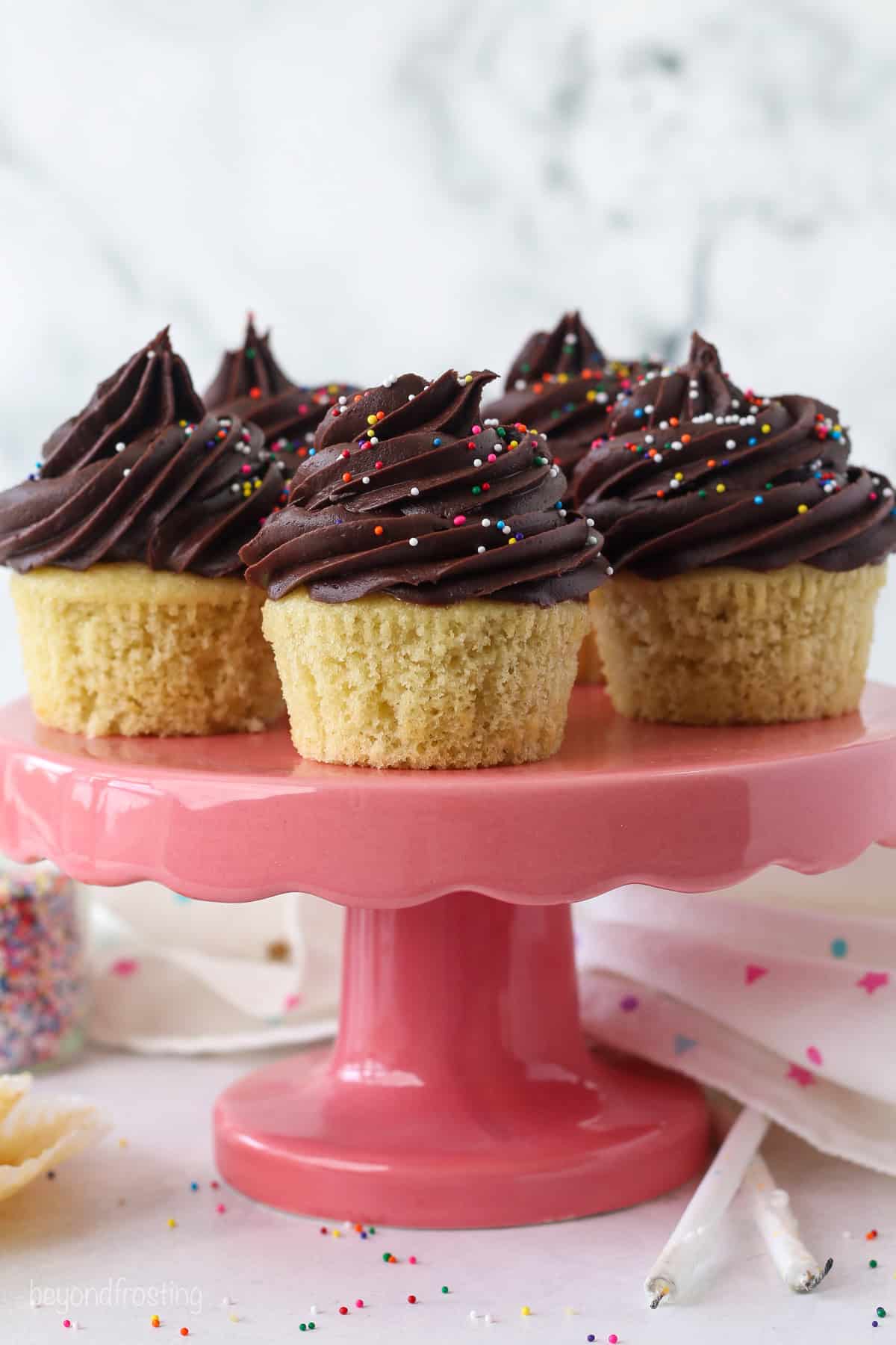 Yellow cupcakes topped with chocolate frosting swirls on a pink cake stand.
