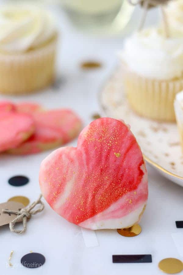A heart shaped sugar cookies with a white and red marble swirled frosting and the cookies are dusted with edible gold glitter. These engagement themed cookies are perfect!