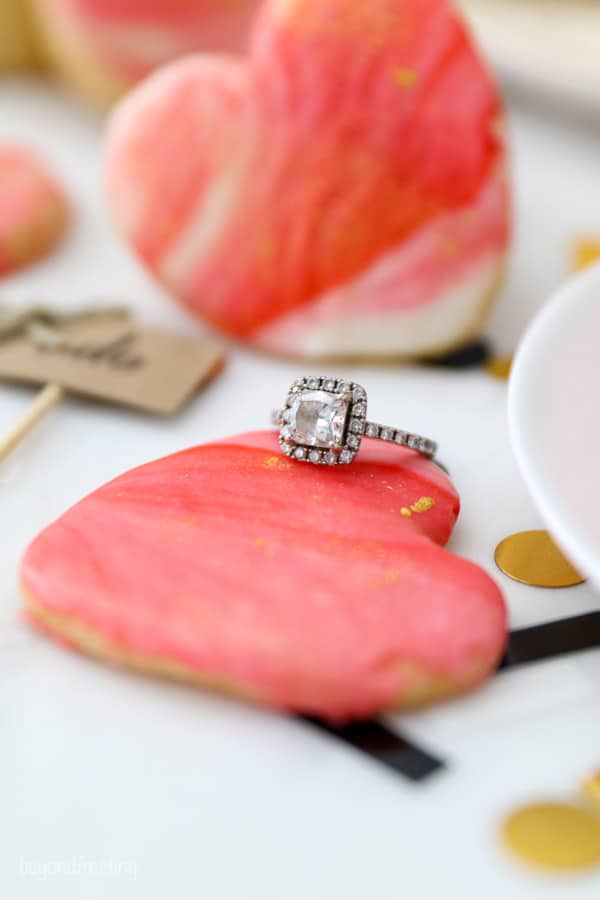 A gorgeous, princess cut diamond engagement ring leaning up against a heart shaped cookie dusted with gold edible glitter