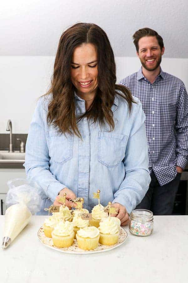 Husband and wife engagement photos in their home kitchen with a plate of cupcakes, a piping bag and sprinkles. The bride is garnishing the cupcakes and the groom is blurred out in the background smiling.