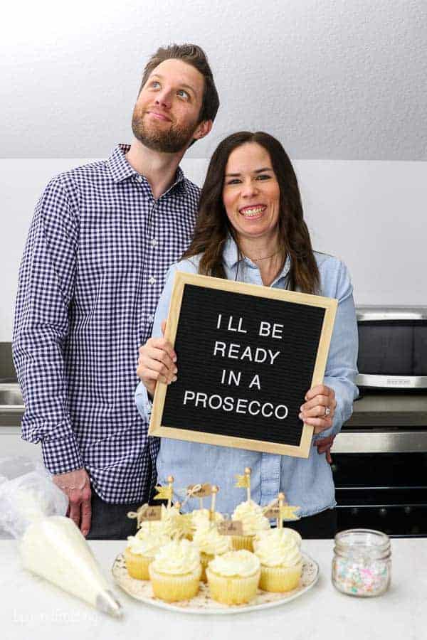 Husband and wife engagement photos in their home kitchen with a plate of cupcakes, a piping bag and sprinkles. The bride is holding a letterboard sign that says "I'll be ready in a prosecco"