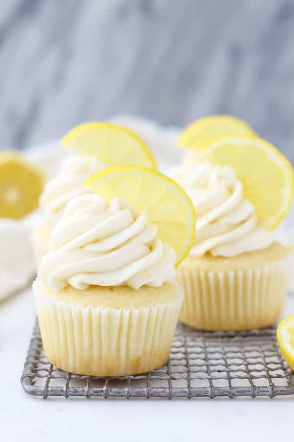 A gorgeous lemon cupcake sitting on a wire rack with a sliced of lemon for garnish on top.