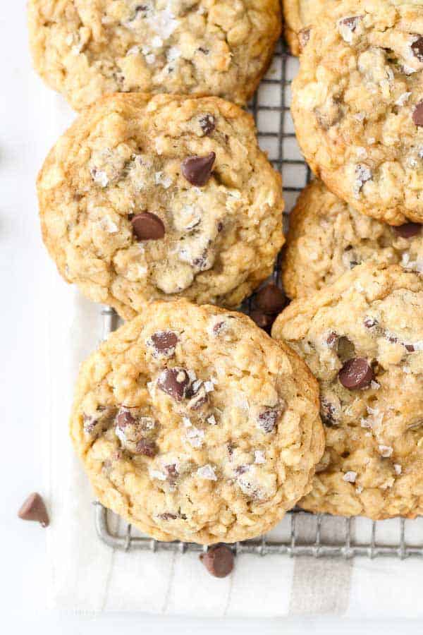 A close up of oatmeal chocolate chip cookies with large flakes of seas salt on top are laying on a vintage wire cooling rack