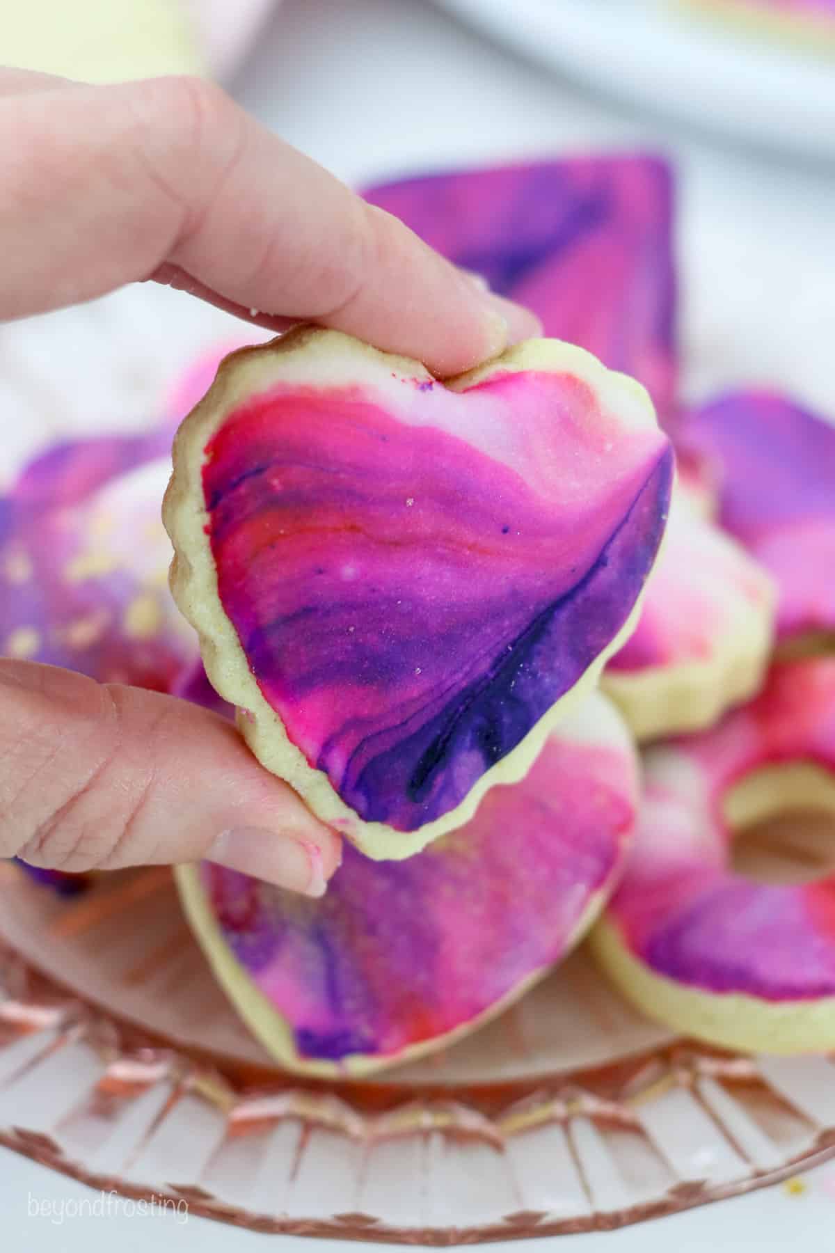 A hand holding an iced heart-shaped cookie over more Valentine's Day cookies on a plate.