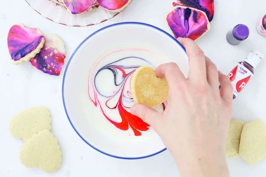 A hand is dunking a cookie into white icing that is swirled with different gel colors