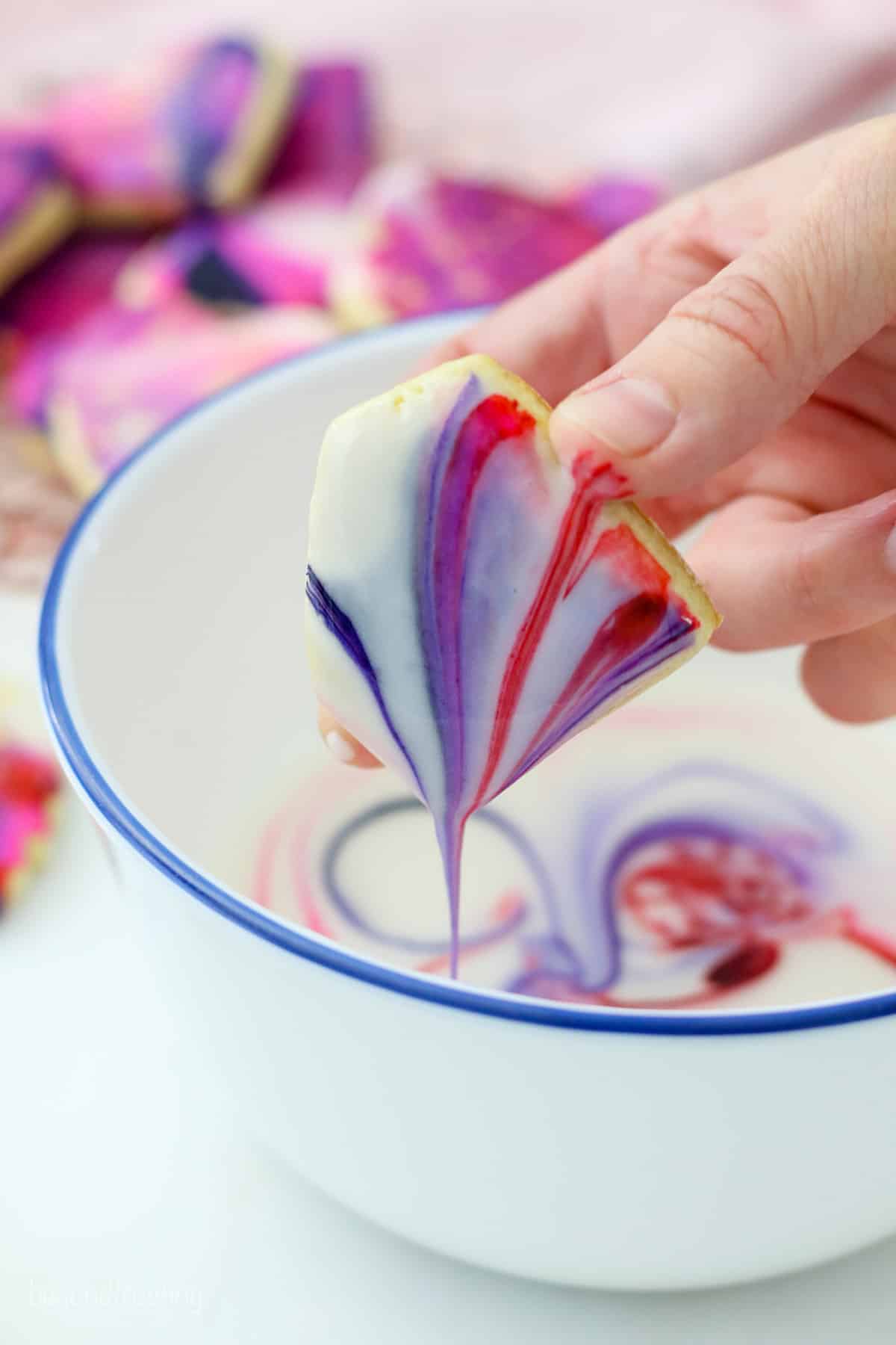 A hand dipping a heart-shaped sugar cookie into a bowl of marbled icing.