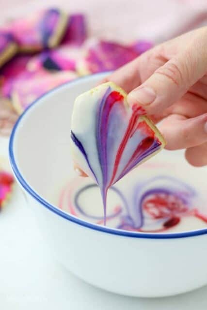 A hand is holding a diamond shaped sugar cookie that has been dipped in marbled icing. The icing is dripping off the cookie back into the bowl showing the beautiful design.