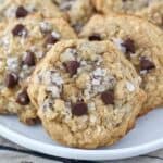 A white rimmed plate is stacked full of chunky oatmeal chocolate chip cookies with flaky seat salt on top