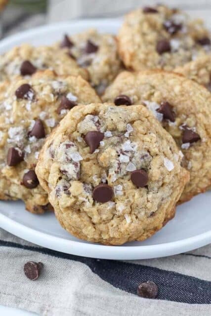 A white rimmed plate is stacked full of chunky oatmeal chocolate chip cookies with flaky seat salt on top