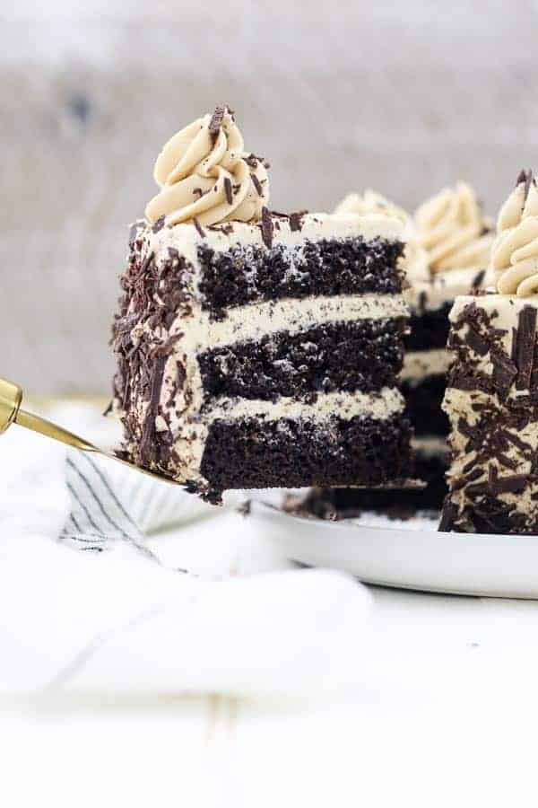 A gold serving spatula is holding a slice of 3 layer of chocolate cake with mocha buttercream and chocolate shavings.