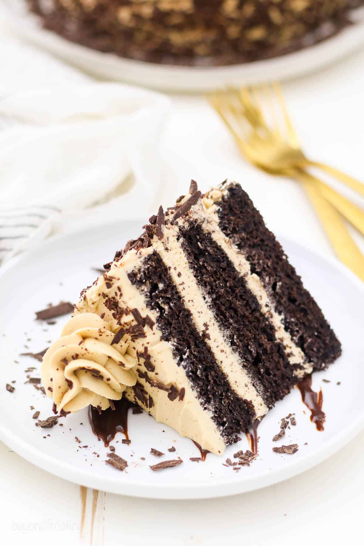 A slice of frosted 3-layer chocolate mocha cake laying on its side on a white plate.