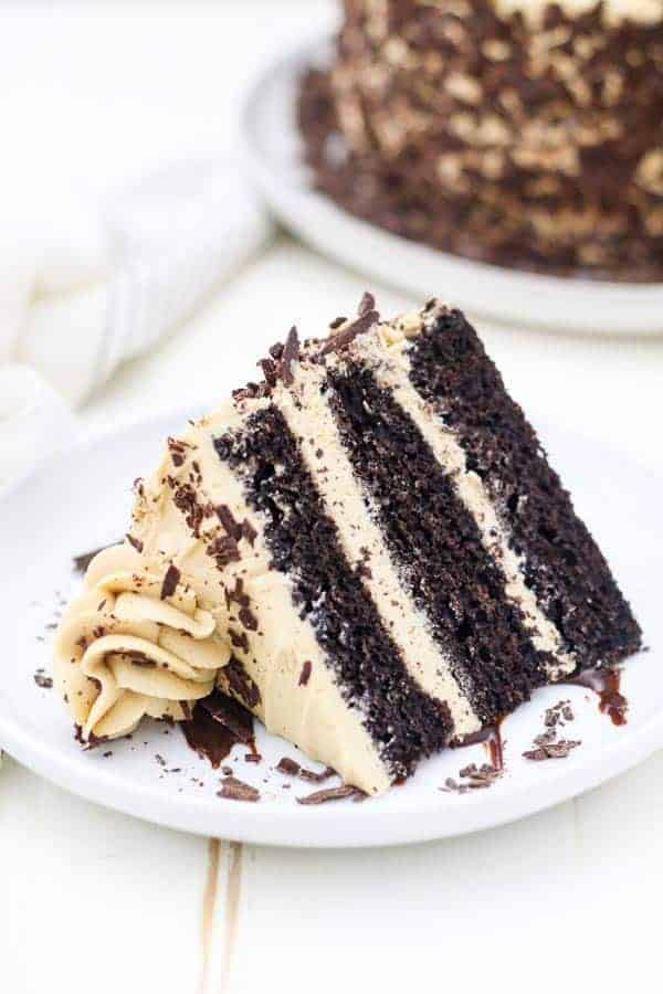 A 3 layer chocolate cake covered in a mocha buttercream is laying sideways on a white rimmed plate and the larger cake is blurred out in the background