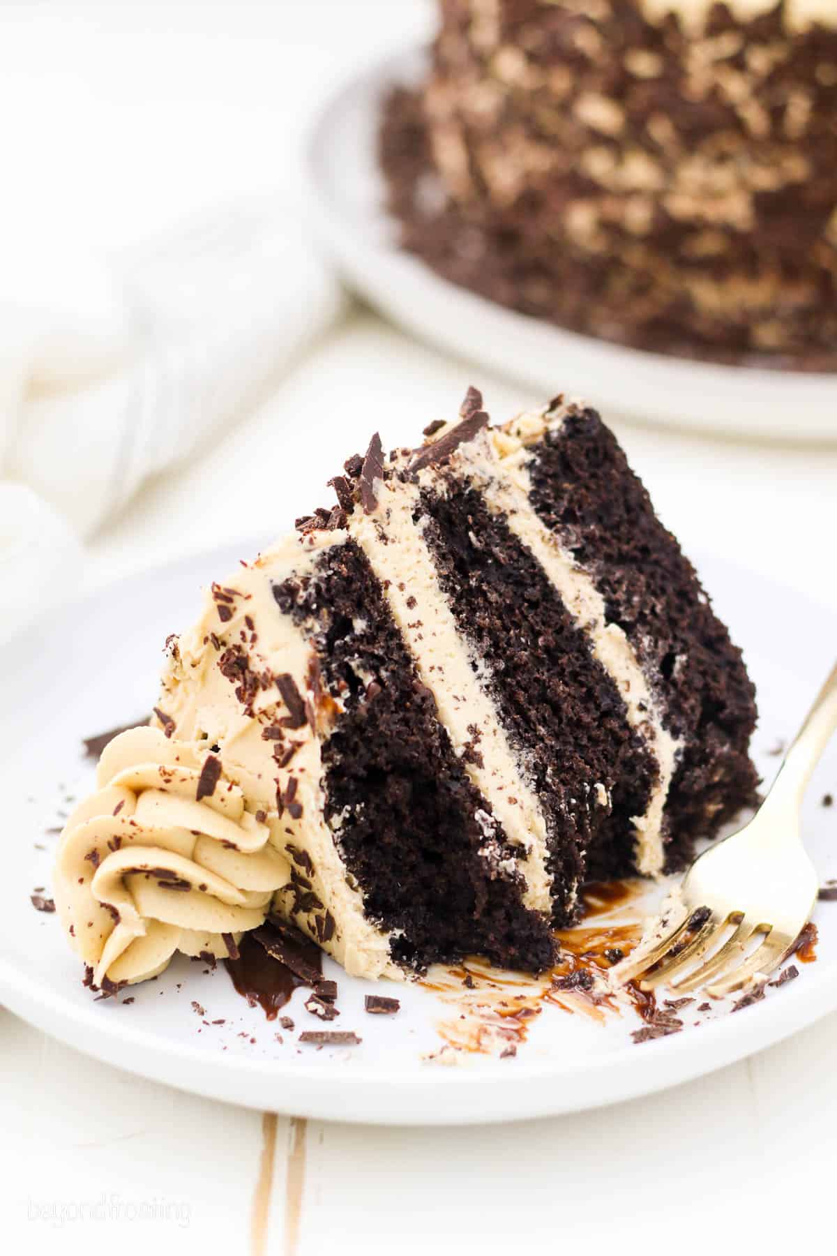 A slice of frosted 3-layer chocolate mocha cake laying on its side on a white plate, with a corner missing.