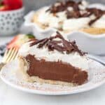 A horizontal images of a slice of chocolate pie with whipped cream sitting on a white plate that has been dusted with cocoa powder. In the background you can see the fill sized pie and several strawberries.