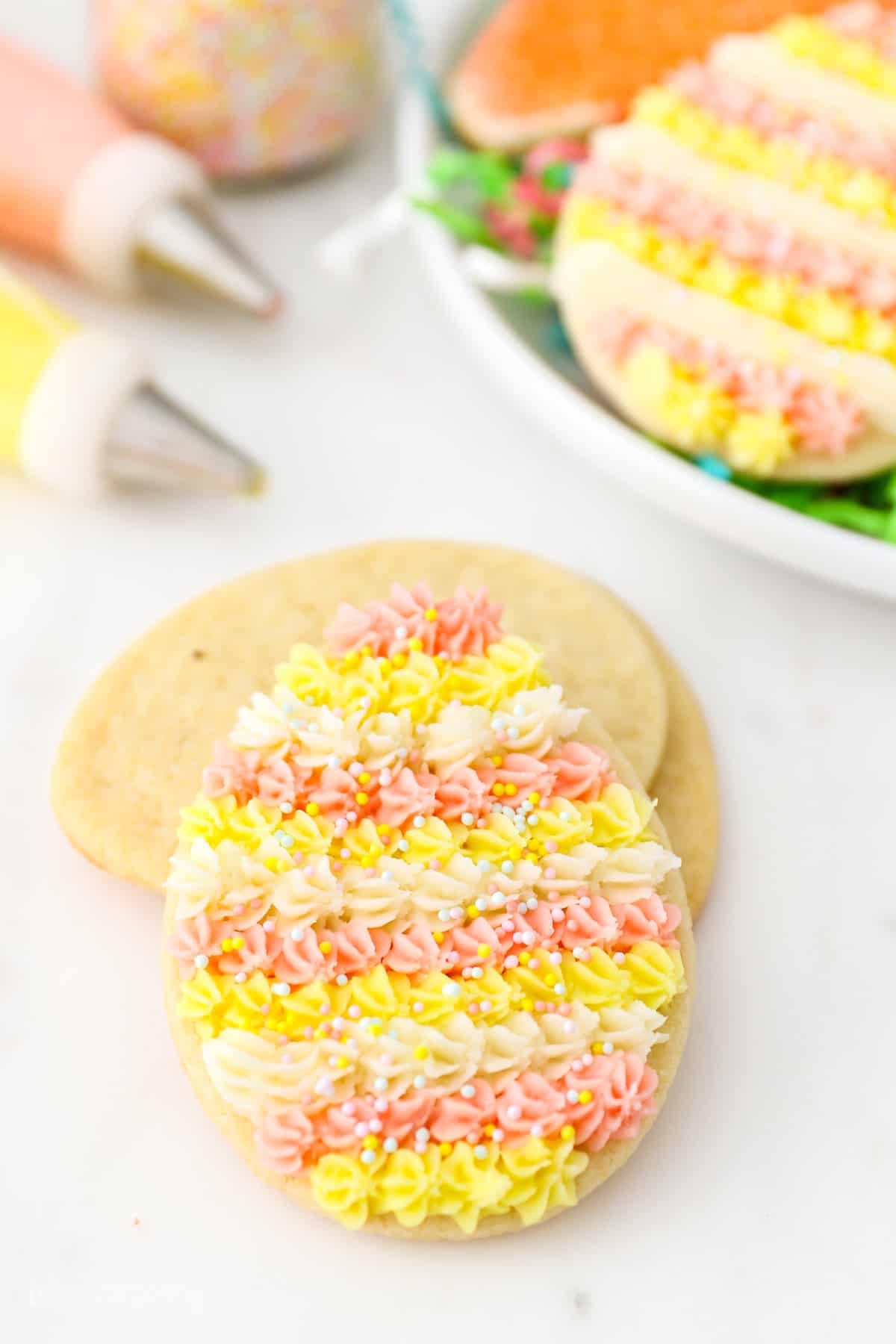 A frosted Easter egg cookie leaning against a stack of unfrosted sugar cookies, with more cookies on a plate next to piping bags in the background.