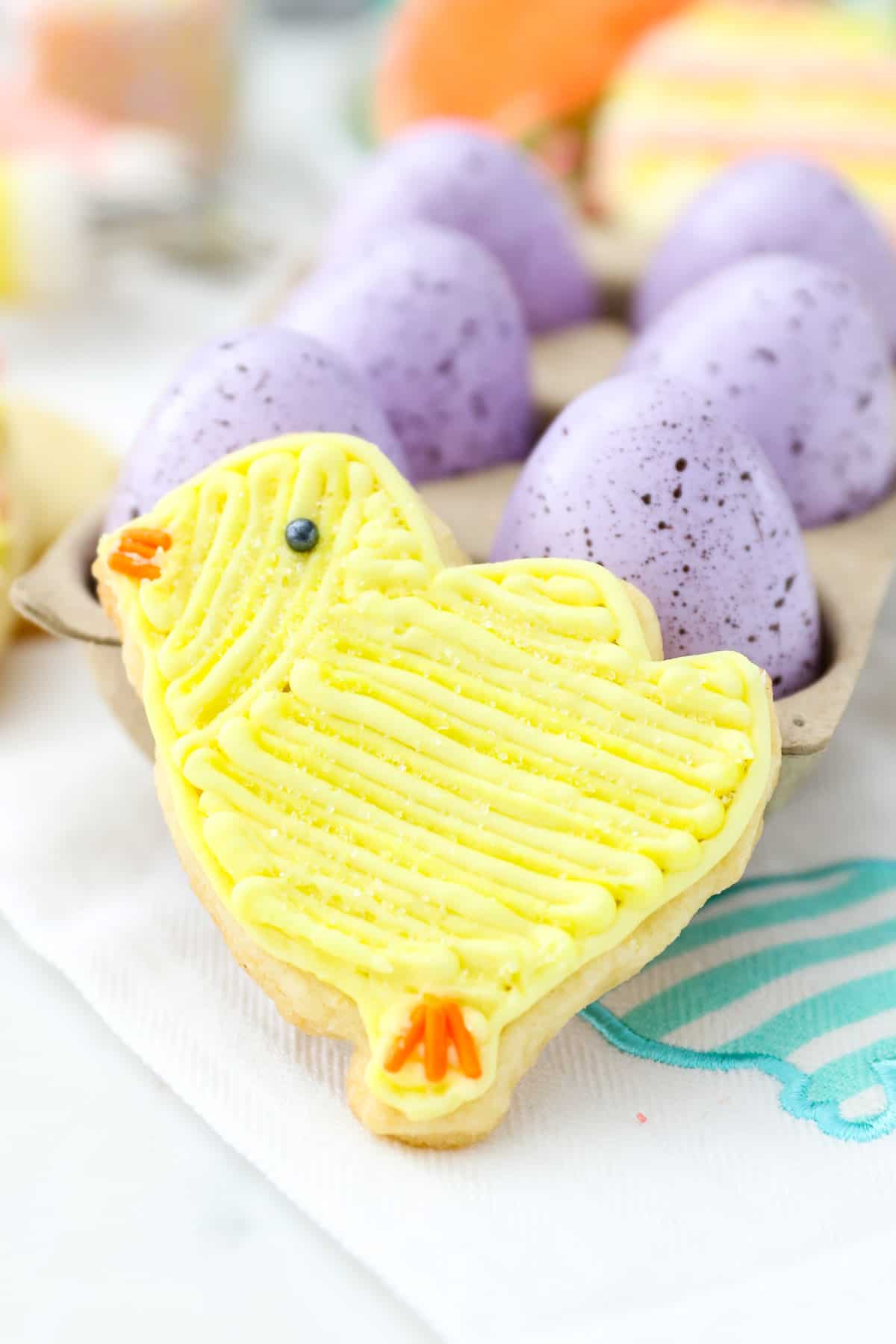 A chick-shaped Easter cookie frosted with yellow buttercream frosting, propped up against a carton of speckled Easter eggs.