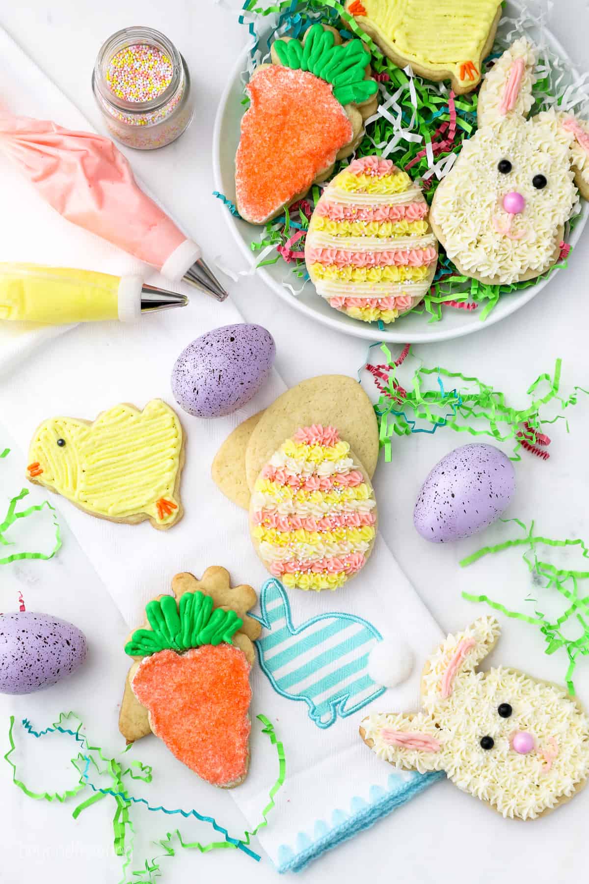 Overhead view of Easter sugar cookies on a plate, next to more assorted Easter cookies scattered on a white countertop next to speckled eggs and decorating tools.