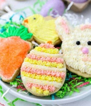 Adorable Easter cookies sitting on paper grass. There's an Easter egg, a bunny face, a carrot and a chick. The cookies are decorated with pink and yellow buttercream and sprinkles.