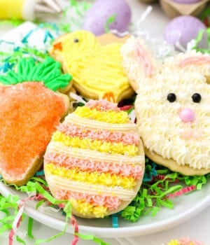 Frosted Easter sugar cookies shaped like a bunny, egg, chick, and carrot on a plate, nestled in green paper grass.