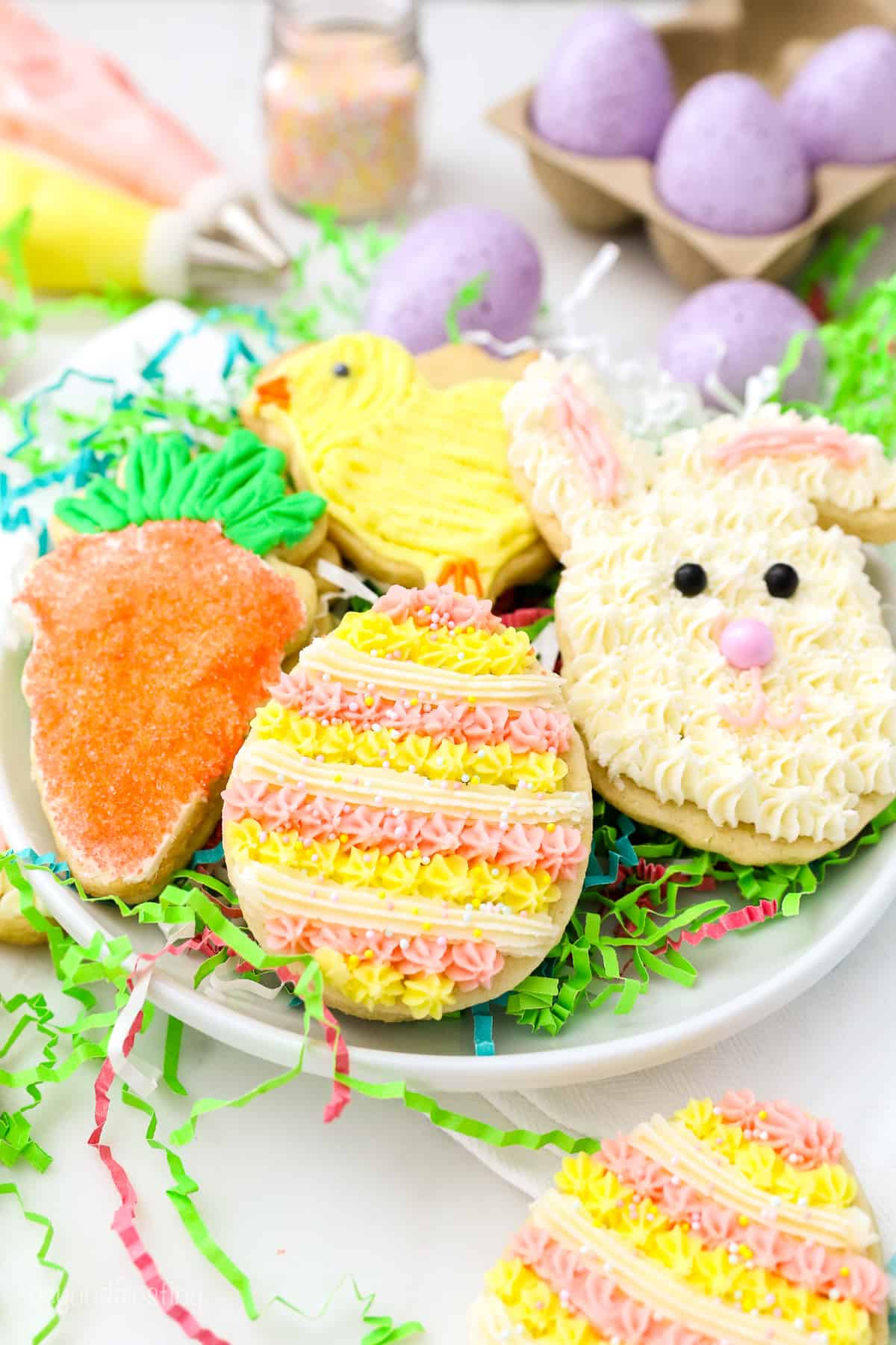 Frosted Easter sugar cookies shaped like a bunny, egg, chick, and carrot on a plate, nestled in green paper grass.