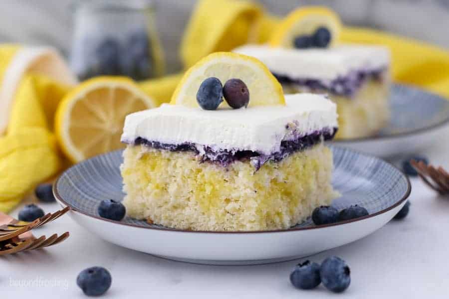 A wide shot of a A close up shot of a slice of lemon blueberry cake, showing the lemon pudding filling, the blueberry topping and the whipped cream is garnished with a lemon slice and a couple of blueberries. The background is blurred out with another slice of cake, a sliced lemon and a jar of blueberries.