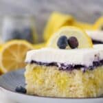 A close up shot of a slice of lemon blueberry cake, showing the lemon pudding filling, the blueberry topping and the whipped cream is garnished with a lemon slice and a couple of blueberries