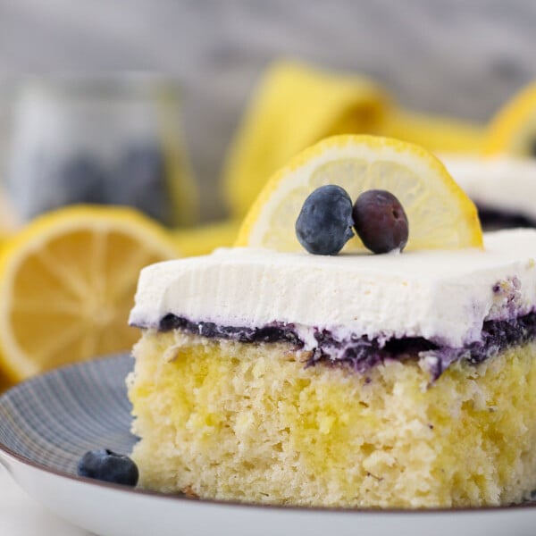 A close up shot of a slice of lemon blueberry cake, showing the lemon pudding filling, the blueberry topping and the whipped cream is garnished with a lemon slice and a couple of blueberries