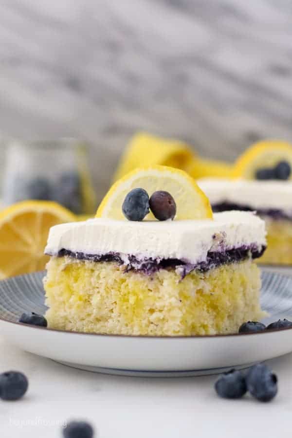 A shot of a slice of lemon blueberry cake, showing the lemon pudding filling, the blueberry topping and the whipped cream is garnished with a lemon slice and a couple of blueberries. There's a lemon and jar of blueberries blurred out in the background