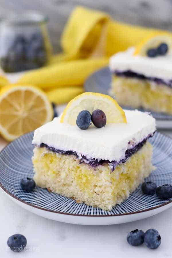 A top down view of a slice of lemon blueberry cake on a blue stripped plate. The slice of cake is garnish with a lemon slice and blueberries.