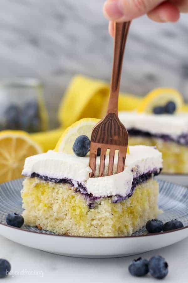 A gold fork is sinking into a a slice of lemon blueberry cake. The slice of cake is sitting on a blue striped plate and it's garnished with blueberries and a sliced lemon