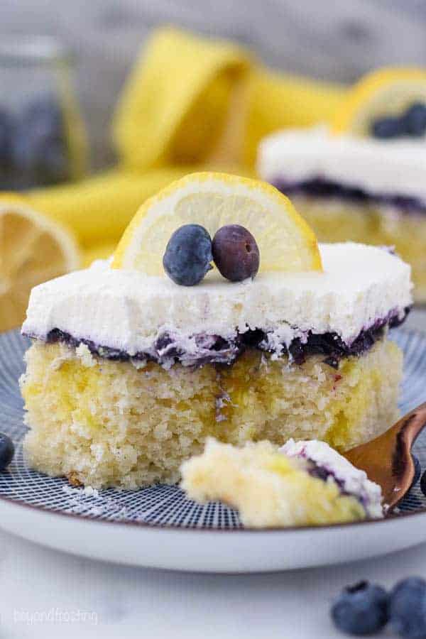 A slice of lemon blueberry cake on a white and blue patterned plate. There's a couple bites missing out of the cake revealing the inside of the cake and the pudding filling.