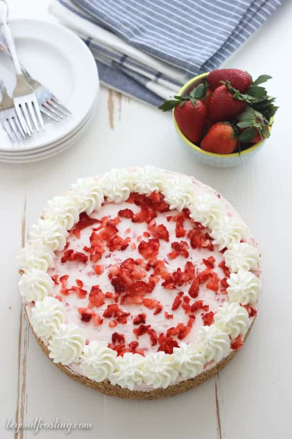 An overhead shot of a strawberry pie with a blue bowl of strawberries in it, a blue napkin in the background and a stack of white rimmed plates with 3 silver forks on top.