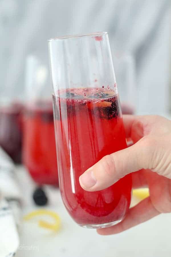 A hand holding a flute glass with a blackberry champagne mixed drink inside