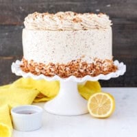 Frosted lemon coconut cake decorated with a skirt of toasted coconut on a white cake stand, on a countertop next to two lemon halves.