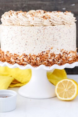 Frosted lemon coconut cake decorated with a skirt of toasted coconut on a white cake stand, on a countertop next to two lemon halves.