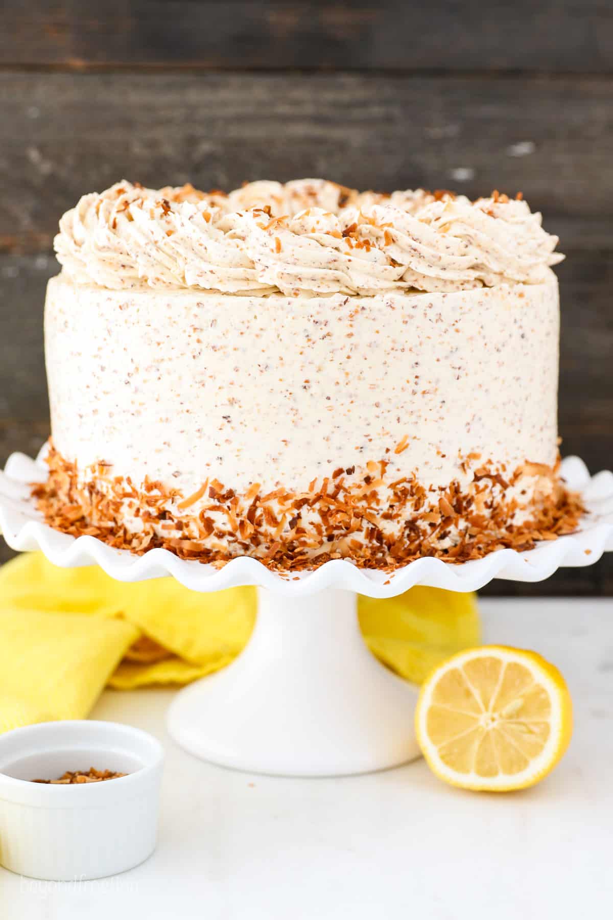 Frosted lemon coconut cake decorated with a skirt of toasted coconut on a white cake stand, on a countertop next to two lemon halves.
