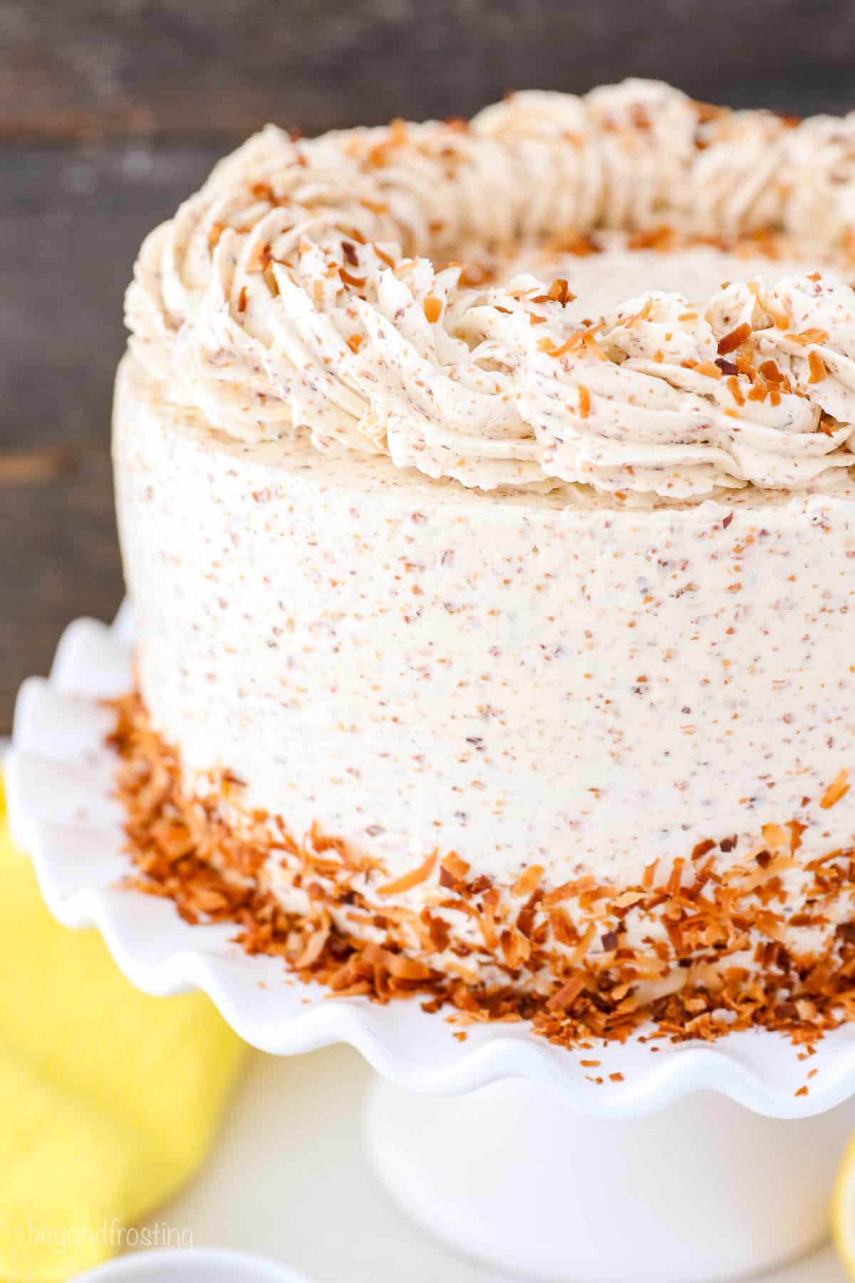 Close up of a frosted lemon coconut cake topped with swirls of frosting, with a skirt of toasted coconut on a cake stand.