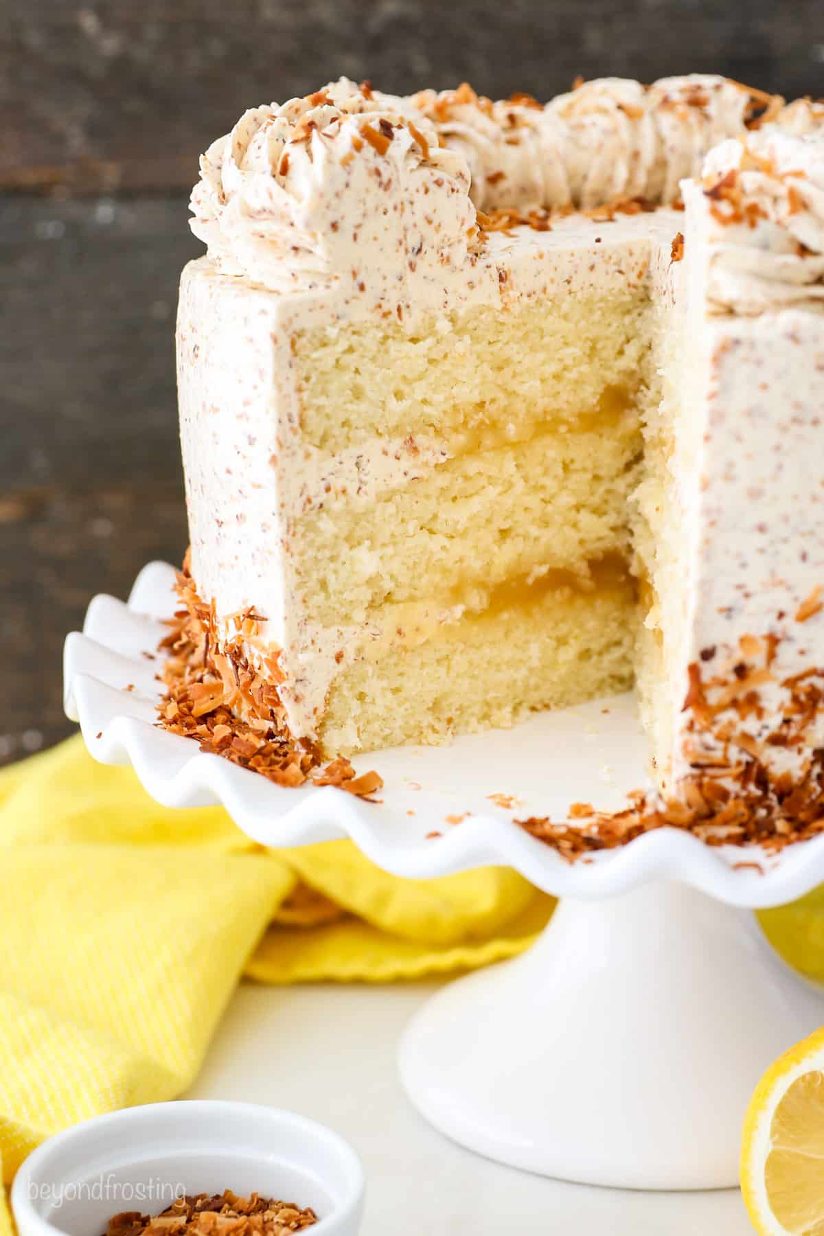 Close up of a frosted lemon coconut cake topped with swirls of frosting, with a skirt of toasted coconut on a cake stand, with a large slice missing.